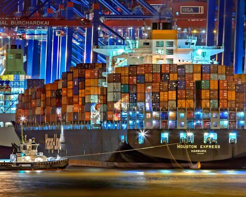 pexels-photo-753331-753331 A brightly lit cargo ship at Hamburg harbor with stacked containers and a tugboat.
